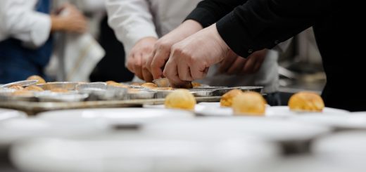 Multiple hands in white coats and black sleeves place small baked buns onto aluminum trays in a busy kitchen prep line in a row.
