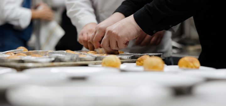 Multiple hands in white coats and black sleeves place small baked buns onto aluminum trays in a busy kitchen prep line in a row.