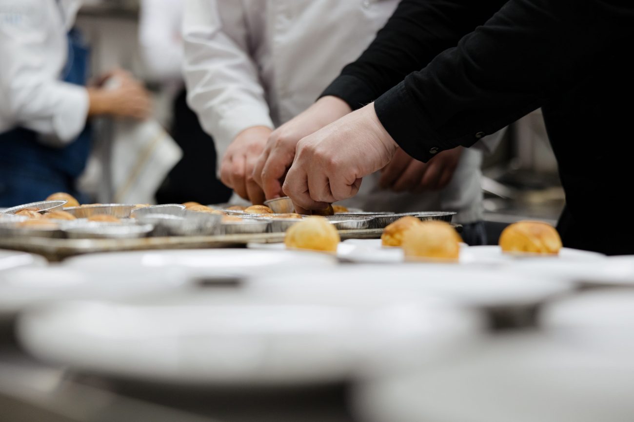 Multiple hands in white coats and black sleeves place small baked buns onto aluminum trays in a busy kitchen prep line in a row.