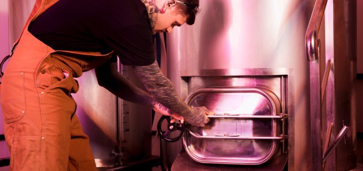 A worker in brown overalls opens a large stainless-steel industrial oven with a circular door in a pink-lit facility.