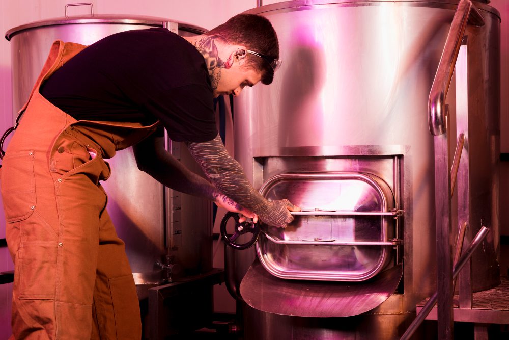 778477 - Multifiltra | Negocios10.Com A worker in brown overalls opens a large stainless-steel industrial oven with a circular door in a pink-lit facility.
