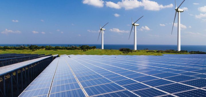 Rows of solar panels in the foreground, wind turbines on a hill beyond, with the ocean in the distance.