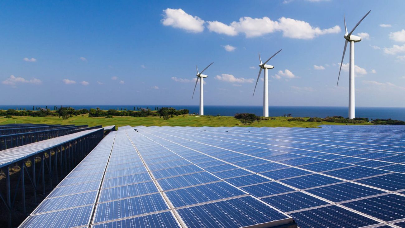 Rows of solar panels in the foreground, wind turbines on a hill beyond, with the ocean in the distance.