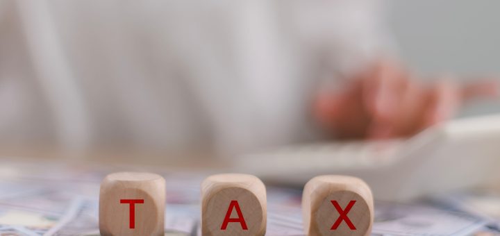 Three wooden letter blocks spell TAX on a surface scattered with dollar bills, conveying the idea of taxes or taxation.