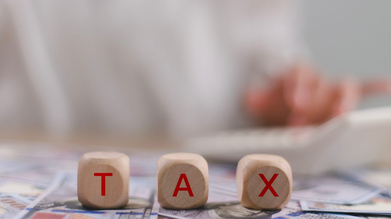 Tax word on wooden cubes with a woman working in the background | Negocios10.Com Three wooden letter blocks spell TAX on a surface scattered with dollar bills, conveying the idea of taxes or taxation.