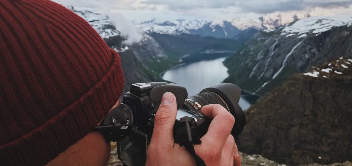 Person wearing a red knit beanie takes a landscape photo with a DSLR over a rocky, snow-capped mountain valley and lake below.
