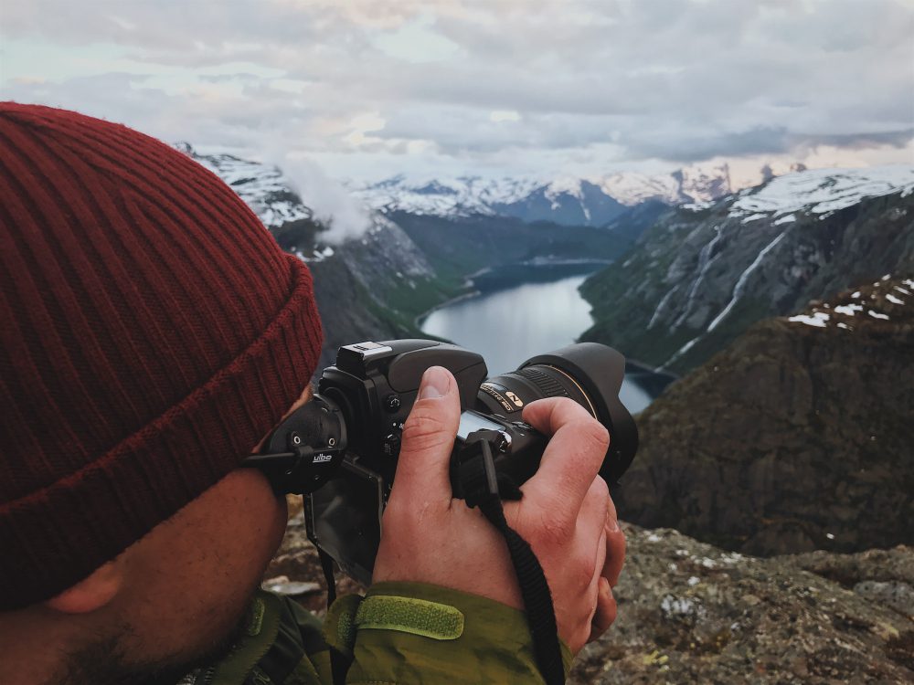 779030 - Coconut Travel | Negocios10.Com Person wearing a red knit beanie takes a landscape photo with a DSLR over a rocky, snow-capped mountain valley and lake below.