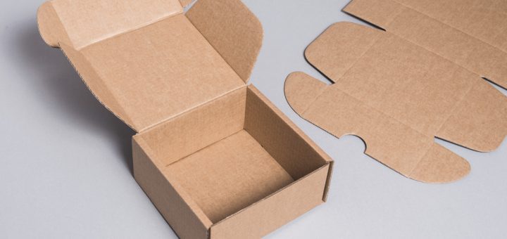 Open brown cardboard box with flaps lifted, sitting on a gray surface, with flat folding carton pieces nearby.