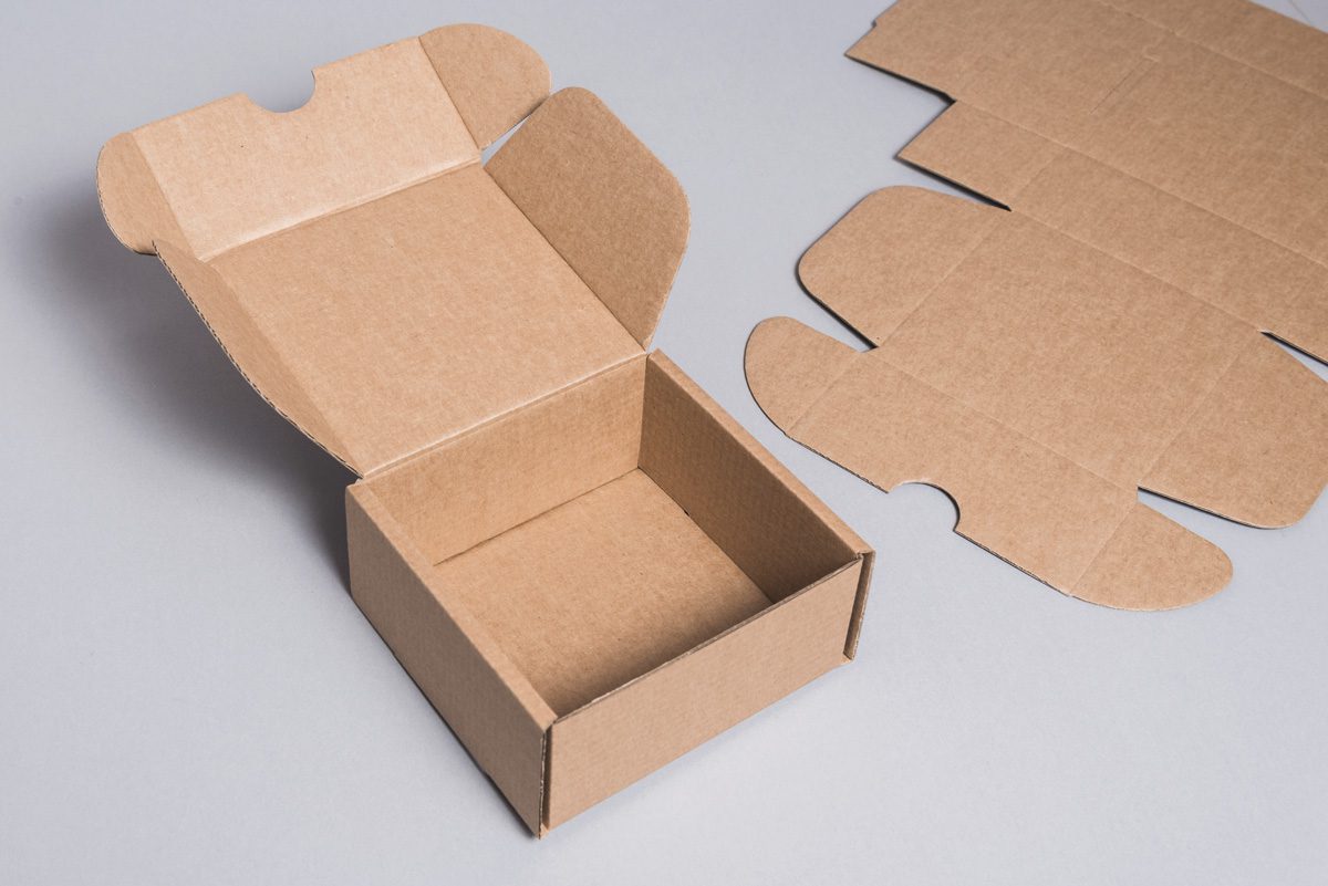 Open brown cardboard box with flaps lifted, sitting on a gray surface, with flat folding carton pieces nearby.