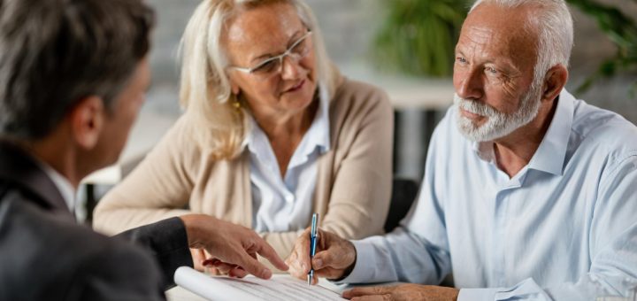 Older man signs a document as a woman looks on with a smile at a table during a business meeting.