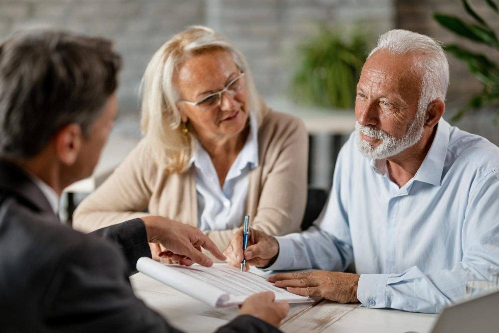 780500 - Devengo Abogados | Negocios10.Com Older man signs a document as a woman looks on with a smile at a table during a business meeting.