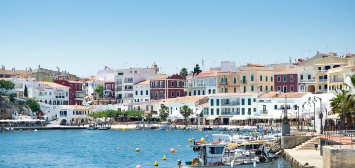 Colorful coastal town with pastel buildings along a calm harbor, with moored boats and a bright blue sky.