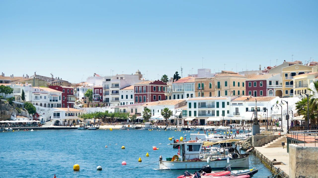 Colorful coastal town with pastel buildings along a calm harbor, with moored boats and a bright blue sky.