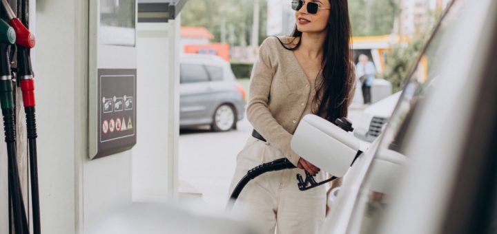 Woman wearing sunglasses at a gas station, holding a fuel nozzle beside a white car.