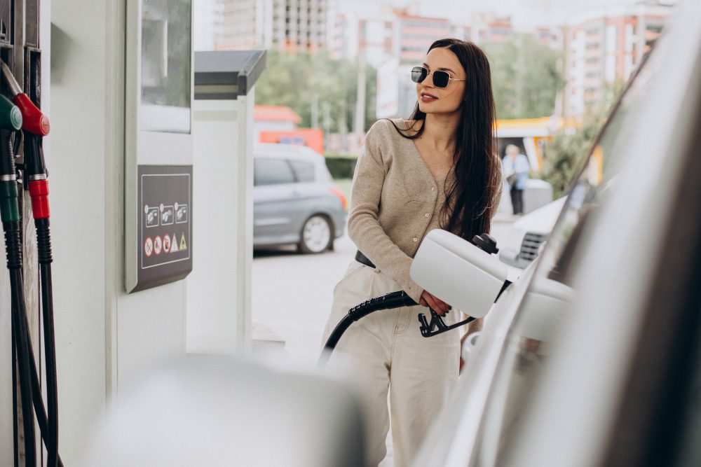 Woman wearing sunglasses at a gas station, holding a fuel nozzle beside a white car.