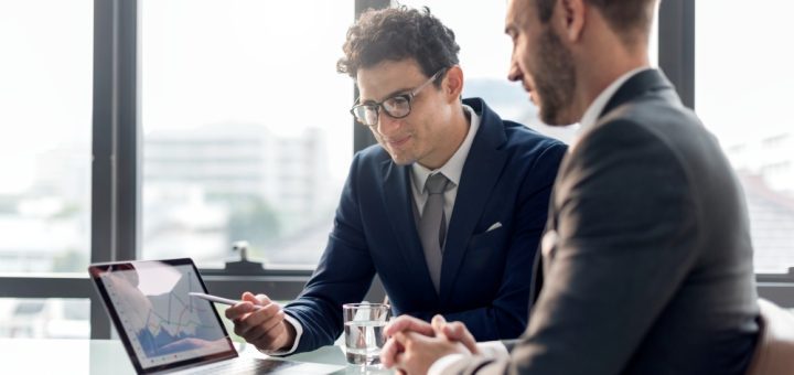 Two businessmen in suits discuss data displayed on a laptop with charts at a glass table by a bright window.