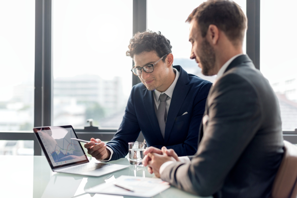 Two businessmen in suits discuss data displayed on a laptop with charts at a glass table by a bright window.