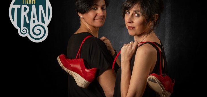 Two women in black, back-to-back, with red sneakers dangling from their shoulders against a dark backdrop; a TRAN TRAN logo appears on the left.