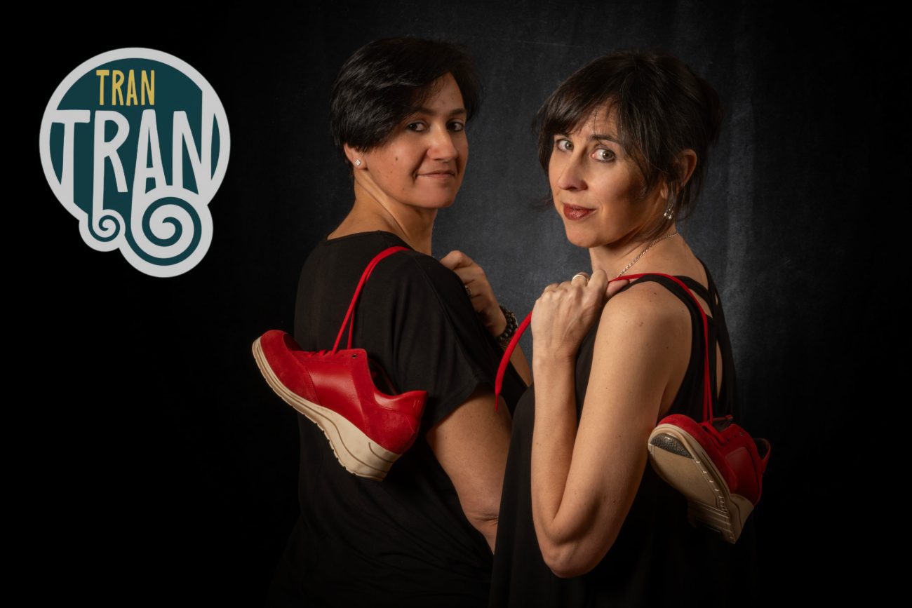 Tran Tran - IP | Negocios10.Com Two women in black, back-to-back, with red sneakers dangling from their shoulders against a dark backdrop; a TRAN TRAN logo appears on the left.