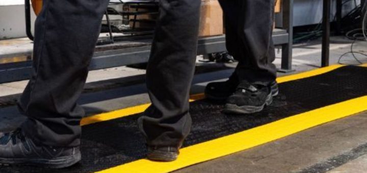 Two workers in black pants and boots stand on a black surface with bright yellow safety stripes on the floor in an industrial setting.