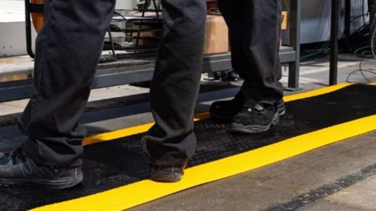 Two workers in black pants and boots stand on a black surface with bright yellow safety stripes on the floor in an industrial setting.